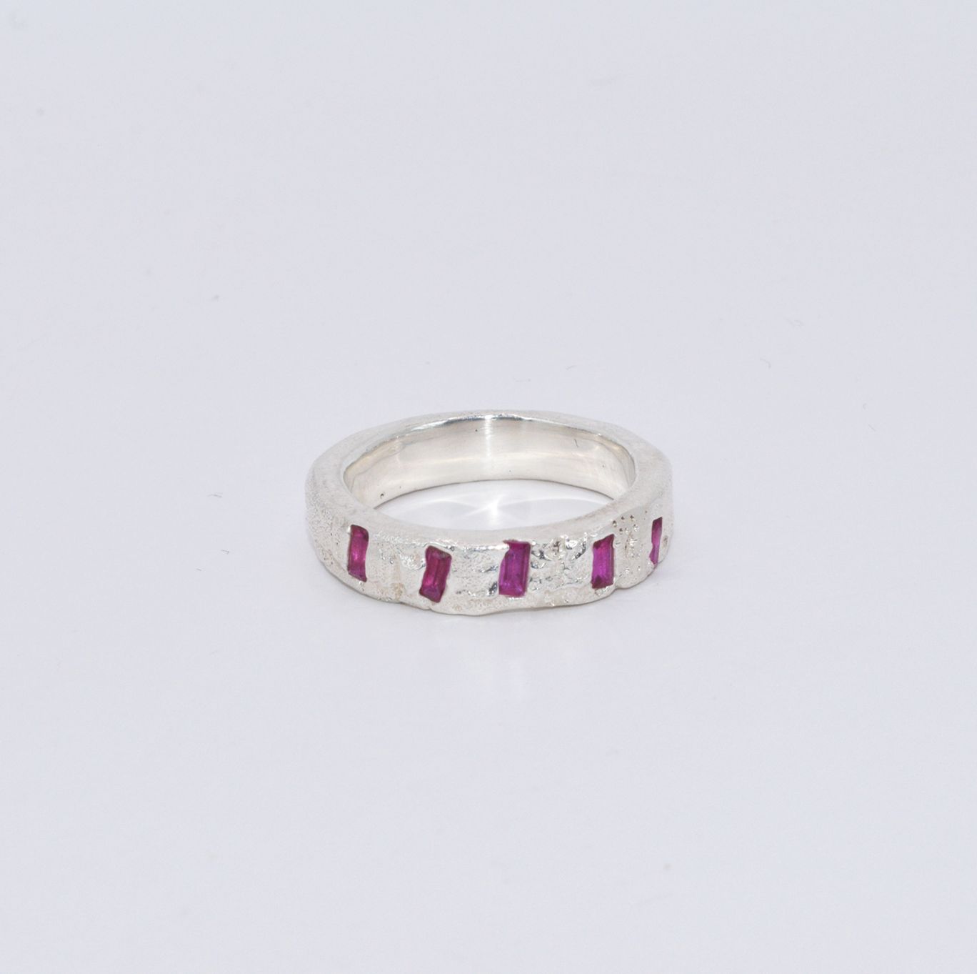Silver ring with five ruby baguettes, arranged in an organic, asymmetrical formation across the band. A raw, textured finish contrasts with the clean geometry of the stones.